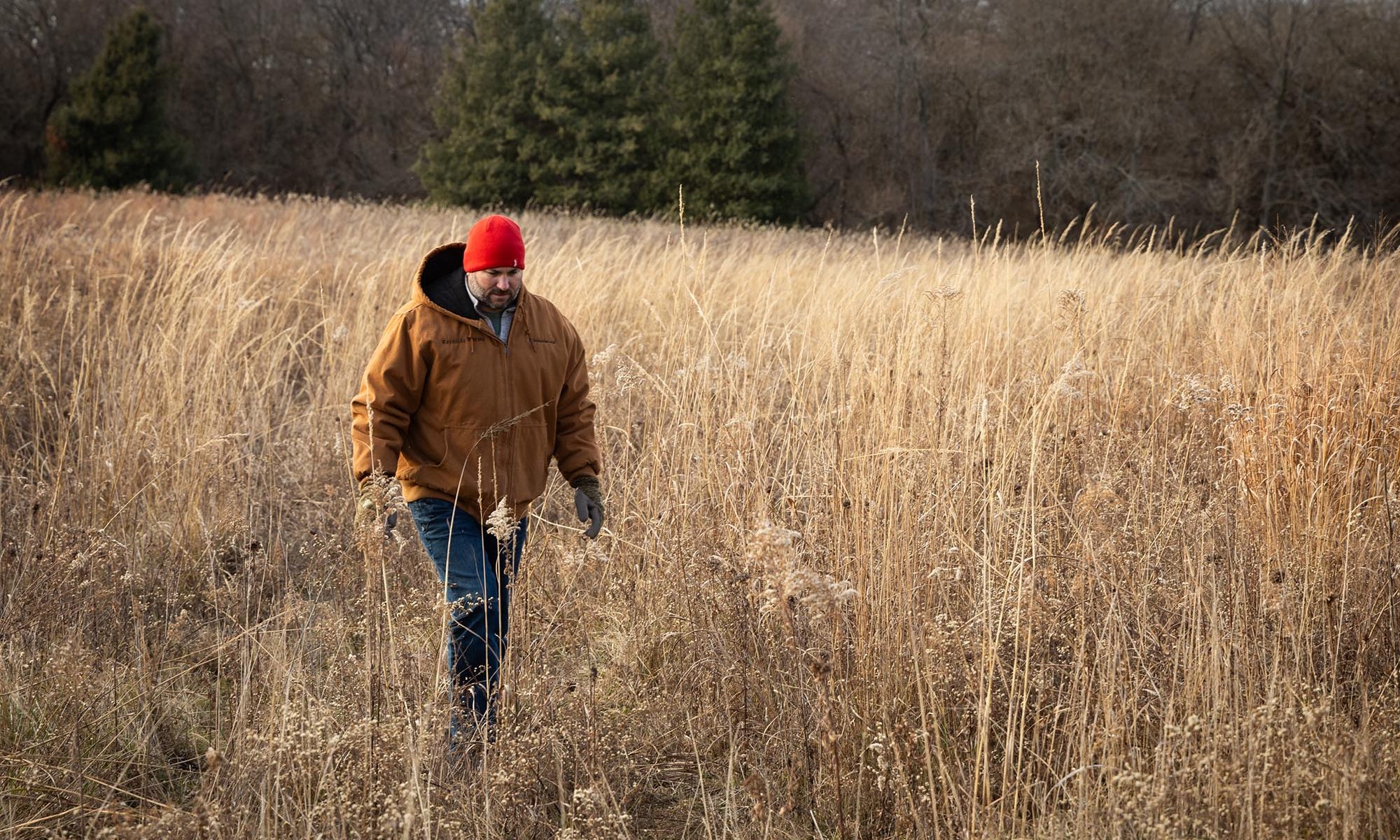 Kris Reynolds wearing red hat and light brown jacket walking in autumnal hued grass.