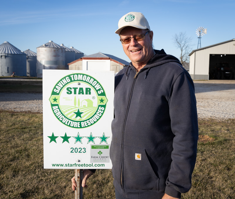 Steve Stierwalt with STAR sign at his farm near Sadorus. Grain bins and machine shed in background.
