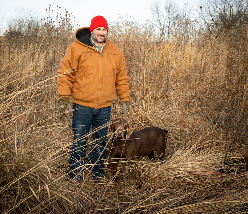 Kris Reynolds in tall native grasses, brown coat and red hat, with hunting dog.