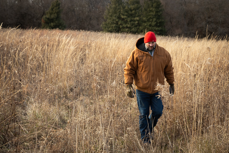 Kris Reynolds wearing red hat and light brown jacket walking in autumnal hued grass.